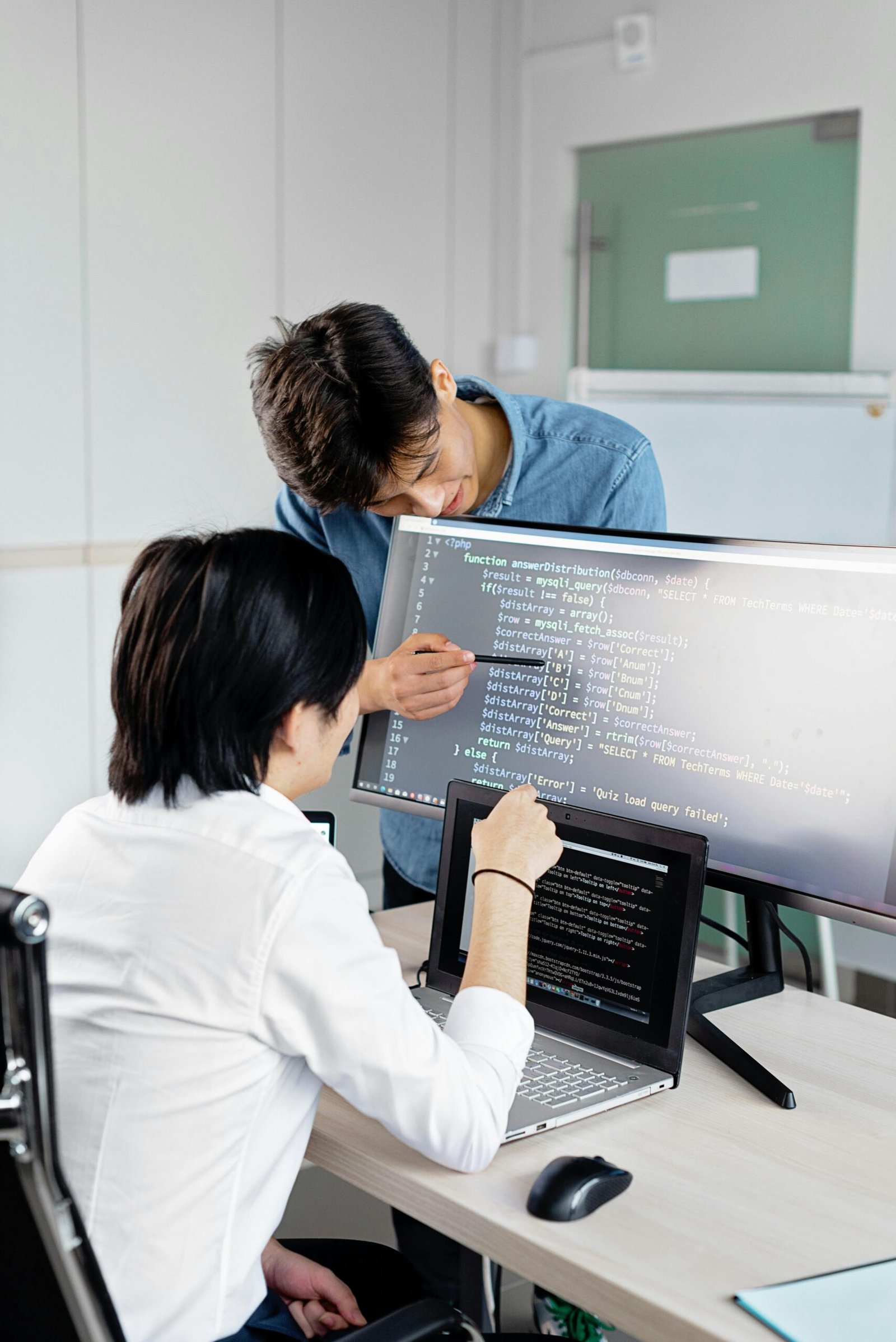 Two professionals collaborating on coding tasks in a modern office environment with laptops and monitors.